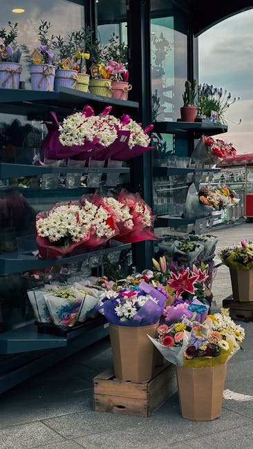 A display of vibrant floral arrangements and bouquets at a flower shop in a glass-fronted setting, with shelves filled with colorful potted plants, wrapped flower bouquets, and decorative pots. Promin